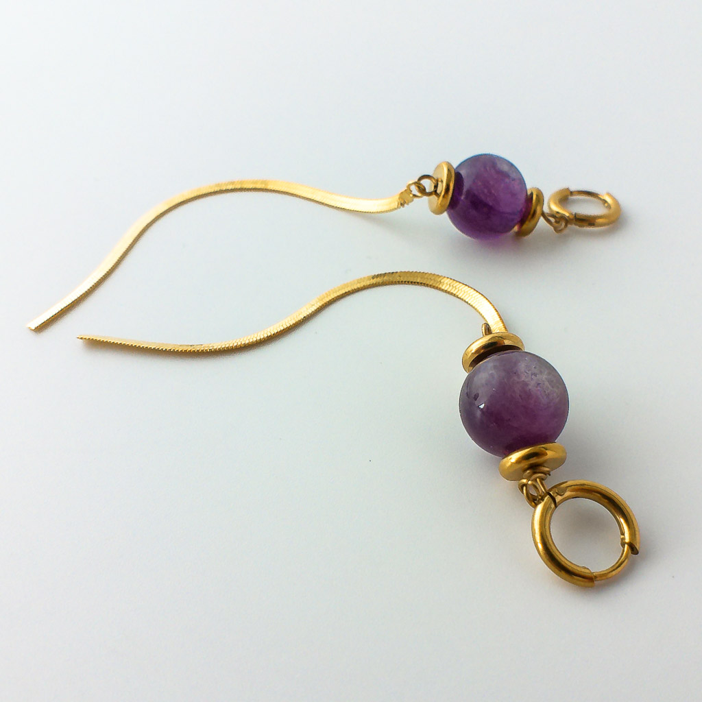 Material: Pendientes de aro de Lepidolita con tira colgante de acero inoxidable. Longitud : 12 cm. Tamaño de la piedra: 1,2 cm. Propiedades de la piedra: Proporciona calma, templanza y confianza en uno mismo. Debido a su alto contenido en litio está indicada contra la tristeza, depresión, ansiedad, miedo y cualquier trastorno psicológico. Ofrece protección y hace salir tu yo superior. Disipa bloqueos. Piedra de transición que libera y reorganiza viejos patrones psicológicos de conducta. Elemento Agua: emociones, deseos, curación, liberación, fluidez, avance. *Las piedras semipreciosas pueden variar ligeramente de tono y forma debido a su origen natural*